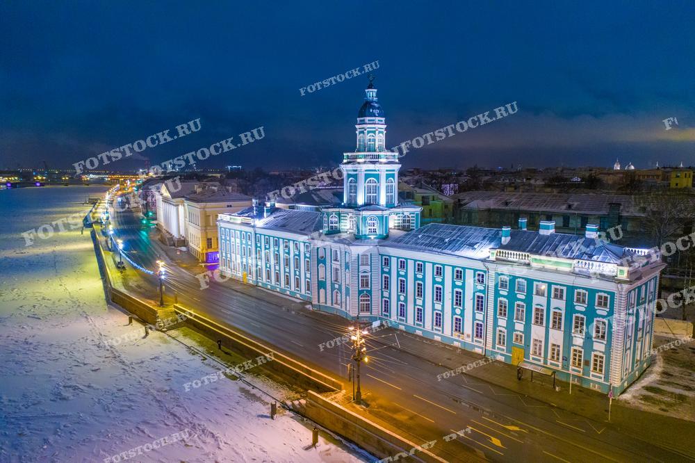 Эта фотография представляет собой вечерний зимний вид на Кунсткамеру в Санкт-Петербурге, расположенную на набережной Невы. Снежное покрывало добавляет к сцене спокойствия и тишины, а освещение здания и улиц подчеркивает архитектурные детали и создает ощущение уюта. Такой снимок будет идеален для использования в качестве иллюстрации в исторических и культурных изданиях, а также как элемент украшения в помещениях.